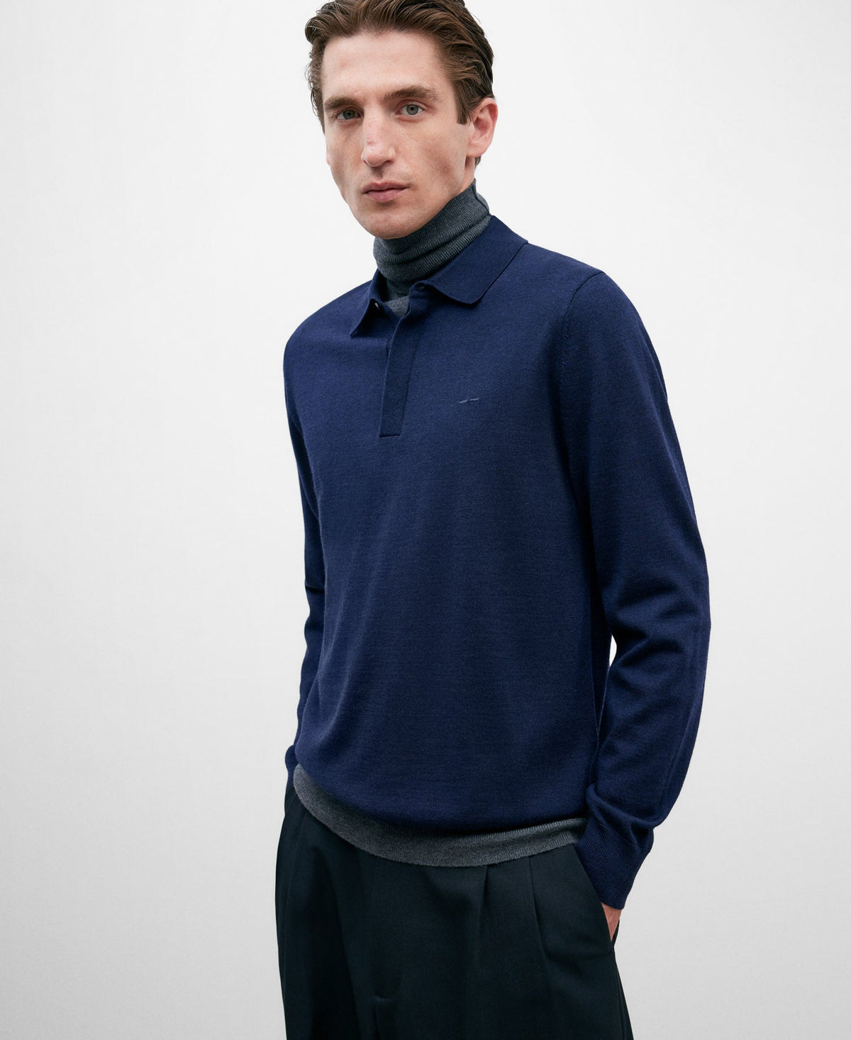 A man wearing a navy blue merino wool long-sleeved polo shirt with a collar and two visible buttons. The shirt features ribbed trims and has an embroidered logo on the chest.